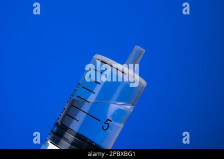 Grande seringue à usage unique sans aiguille à moitié remplie de liquide transparent. Prise de vue en studio macro, isolée sur fond bleu, aucune personne. Banque D'Images