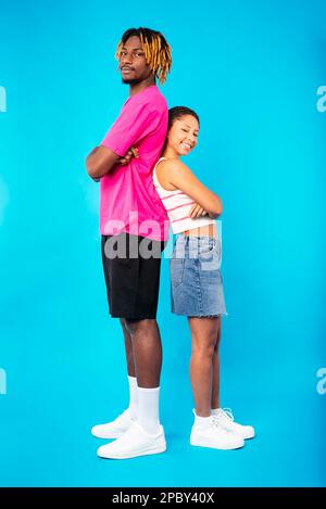 Vue latérale du jeune couple multiethnique bien-aimé dans des vêtements tendance se tenant dos à dos avec des bras croisés et regardant l'appareil photo sur fond bleu Banque D'Images