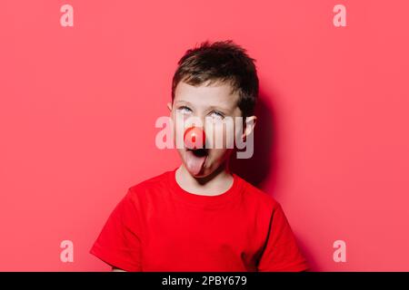 Portrait d'un enfant humoristique faisant un visage drôle avec le nez rouge tout en regardant vers le haut avec l'oeil blanc et la bouche ouverte avec la langue d'arrimage tout en se tenant contre le pois Banque D'Images