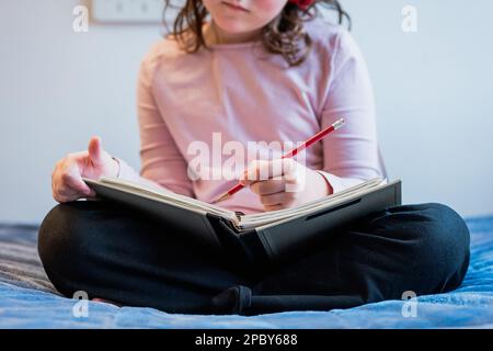 Petite fille de préadolescence ciblée non reconnaissable avec des cheveux bruns ondulés assis sur un lit doux et l'écriture tout en faisant des devoirs dans la chambre à coucher Banque D'Images