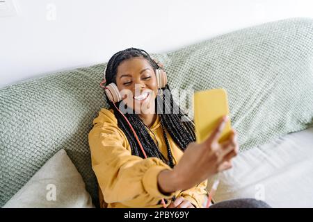 Jeune Afro-américaine positive à capuchon jaune et casque avec tresses souriant et parlant par appel vidéo sur smartphone tout en étant assis sur s. Banque D'Images