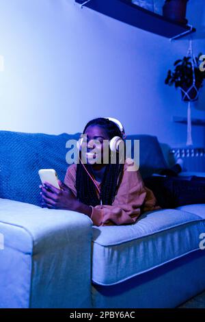 Jeune femme afro-américaine gaie vêtie de vêtements décontractés et écoutez de la musique avec un casque tout en parcourant un téléphone mobile allongé sur un canapé en bleu au néon Banque D'Images