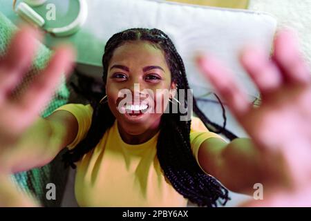 Vue de dessus de jeunes Afro-américaines positives avec des tresses portant un t-shirt jaune souriant et tendant les mains vers l'appareil photo tout en étant allongé sur un canapé Banque D'Images
