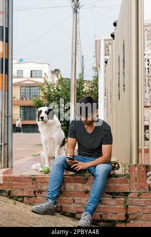 Tout le corps de jeune homme concentré dans des vêtements décontractés assis sur des escaliers de brique et de navigation smartphone près de la jolie frontière Collie contre ville stre Banque D'Images