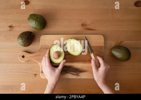 Vue de dessus de récolte anonyme personne avec couteau coupant avocat mûr sur planche à découper sur table en bois intérieure Banque D'Images