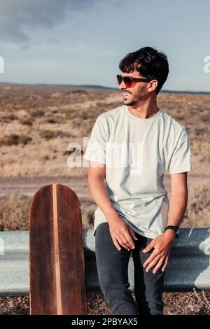 Joyeux jeune homme patineur dans des vêtements décontractés et des lunettes de soleil regardant loin tout en étant assis sur la barrière de bord de route près du long-board contre le terrain sec pendant la pause Banque D'Images