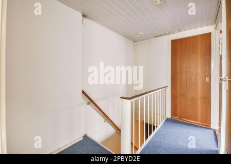 Décoration intérieure de couloir avec murs blancs et porte en bois placée près de l'escalier menant au premier étage avec des rampes et une barrière dans l'appartement Banque D'Images