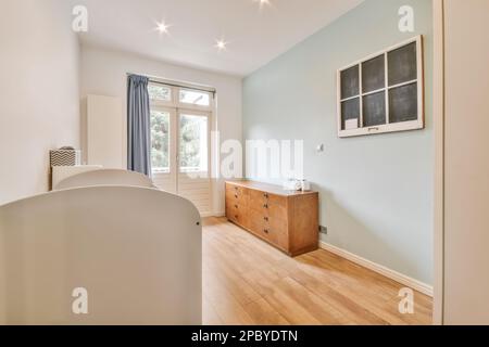 Décoration intérieure de chambre lumineuse avec lit de bébé blanc placé contre une commode en bois près du mur bleu et du balcon Banque D'Images