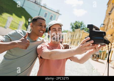 Joyeux homme afro-américain montrant le pouce vers le haut tandis que l'ami prend selfie sur l'appareil photo d'époque sur la descente d'Andrews à Kiev, image de stock Banque D'Images