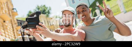 Heureux africain américain montrant le signe de la victoire près de l'ami prenant selfie sur l'appareil photo vintage sur Andrews descente à Kiev, bannière, image de stock Banque D'Images