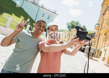 Homme afro-américain souriant montrant le signe de la paix près de l'ami barbu prenant selfie sur l'appareil photo d'époque sur Andrews descente à Kiev, image de stock Banque D'Images