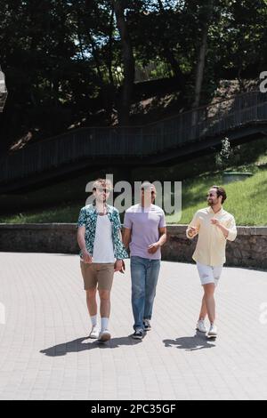 Jeune homme en lunettes de soleil et vêtements décontractés parlant à des amis interraciaux tout en marchant dans le parc, image de stock Banque D'Images