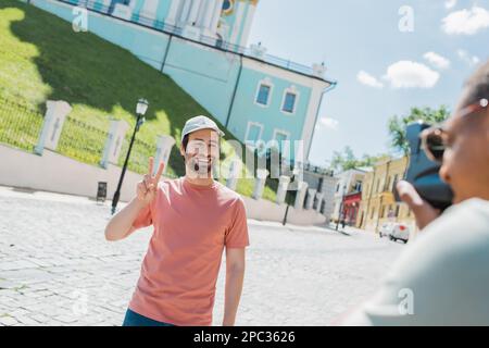 Un homme américain africain flou avec un appareil photo vintage prenant la photo d'un touriste barbu heureux montrant le signe de la victoire sur la descente Andrews à Kiev, image de stock Banque D'Images