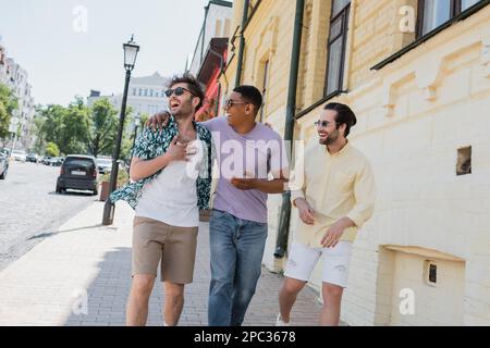 Amis multiethniques positifs dans des lunettes de soleil embrassant et marchant sur Andrews descente à Kiev, image de stock Banque D'Images