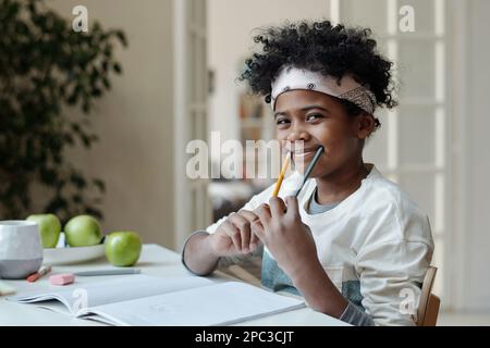Adorable écolier afro-américain s'amusant en s'asseyant au bureau dans la salle de séjour et en tenant deux crayons par les bords de la bouche et en souriant Banque D'Images