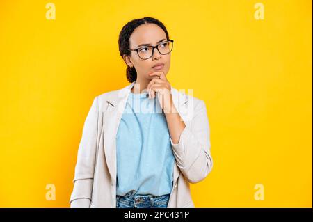 Pensive inquiète hispanique ou brésilienne jeune brune femme d'affaires, avec des lunettes, tient sa main près du menton, regardant de côté, rêvant, planification, penser, se tenir sur fond jaune isolé Banque D'Images