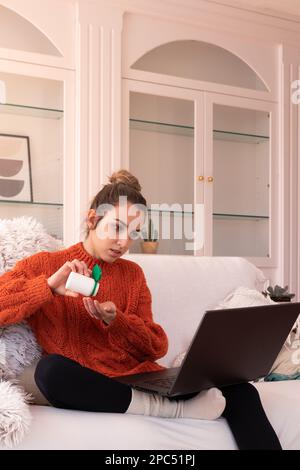 Jeune femme sérieuse dans un chandail chaud assis sur un canapé et prenant des pilules dans un contenant en plastique tout en ayant la conversation en ligne avec le médecin sur l'ordinateur portable du Banque D'Images