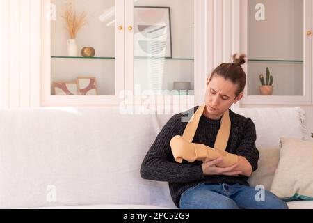 Énervé jeune homme dans des vêtements décontractés avec une main cassée portant un bras de toile assis sur un canapé dans la salle de séjour de l'appartement Banque D'Images