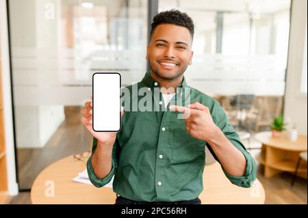 Un homme d'affaires indien souriant tient son smartphone à la main et pointe le doigt sur un écran de téléphone vide, un homme d'étonnement qui présente la meilleure affaire et fait de la publicité pour une nouvelle application mobile au bureau Banque D'Images