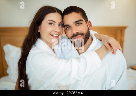 Couple qui s'embrasse en portant des peignoirs blancs qui se posent dans la chambre à l'intérieur Banque D'Images