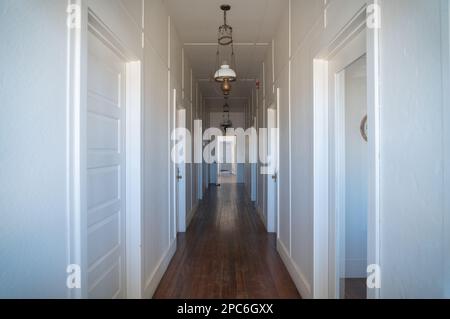 Les portes lambrées et les lampes suspendues du long couloir blanc convergent vers une fenêtre lumineuse au Colonel Allensworth State Historic Park, en Californie. Banque D'Images
