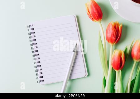 Bloc-notes en spirale, stylo et fleurs de tulipe rouges sur fond vert clair. Vue de dessus, plan d'agencement, espace de copie. Banque D'Images