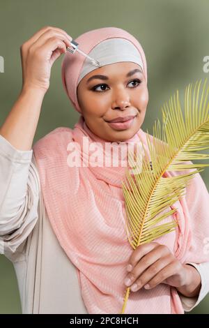 femme musulmane multiraciale dans hijab rose tenant la feuille de plante exotique et appliquant le sérum hydratant sur fond vert, image de stock Banque D'Images