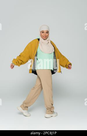 pleine longueur de gaie multiraciale musulmane femme dans blouson blouson jaune et pantalon beige marchant sur fond gris, image de stock Banque D'Images