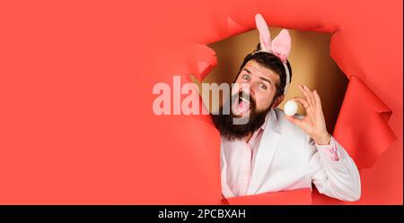 Bonne journée de Pâques. Homme barbu dans les oreilles de lapin avec oeuf blanc regardant à travers le trou de papier. Lapin Bunny homme se préparant pour Pâques. Chasse aux œufs. Pâques Banque D'Images