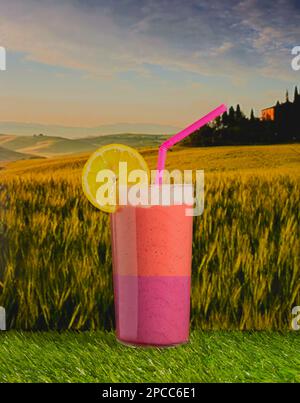 Boisson fouettée colorée garnie d'un segment de citron reposant sur un peu d'herbe, avec des champs en arrière-plan. Banque D'Images