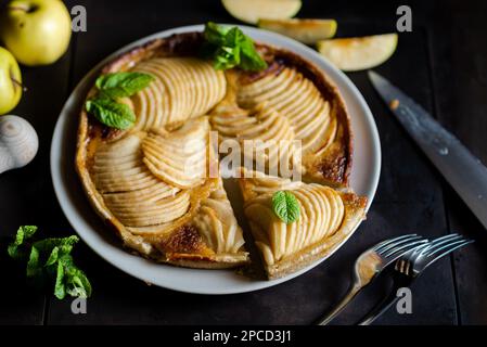 Tarte française aux pommes : tentation sur fond noir. Tarte traditionnelle ouverte avec fines tranches de pommes, décorée de feuilles de menthe. Banque D'Images