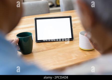 Image rognée d'un couple de personnes âgées biraciales avec des tasses à café en utilisant un comprimé sur une table en bois à la maison Banque D'Images