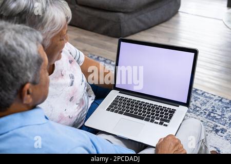 Vue en grand angle du couple biracial senior utilisant un ordinateur portable tout en se relaxant sur le canapé dans le salon Banque D'Images