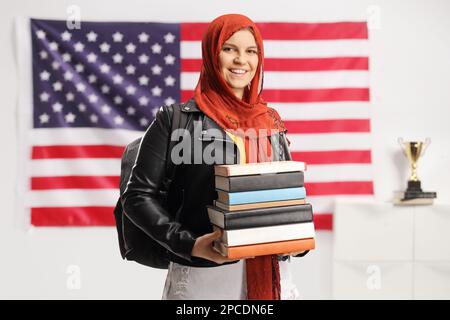 Étudiante musulmane avec hijab portant une pile de livres et souriant devant la caméra devant le drapeau des États-Unis Banque D'Images