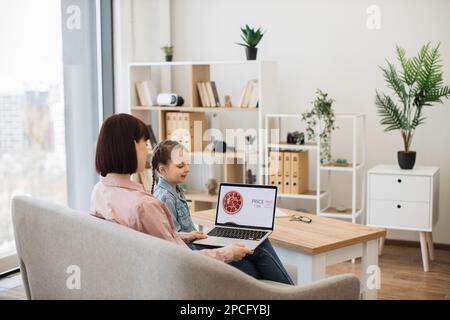 La mère caucasienne aux cheveux foncés et sa belle fille se reposent ensemble dans un salon confortable et utilisent un ordinateur portable moderne pour commander de la nourriture. Joyeuses dames à c Banque D'Images