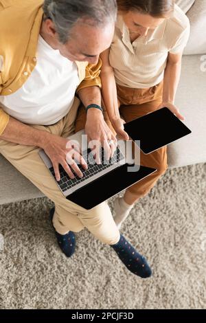 Au-dessus de la vue d'un couple senior assis sur un canapé avec un ordinateur portable et une tablette numérique, maquette pour la conception d'application ou de site Web sur les écrans Banque D'Images