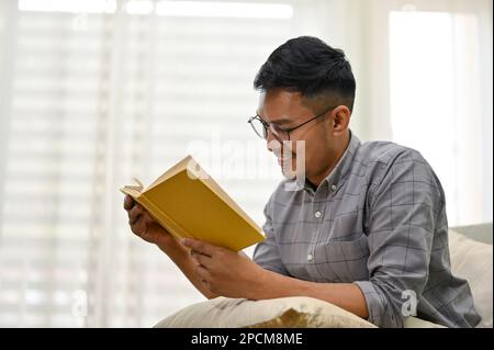 Homme asiatique du millénaire élégant et heureux portant des lunettes lisant un livre sur un canapé dans son salon. vue latérale Banque D'Images