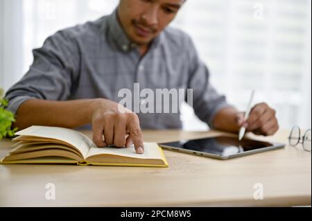 Image rognée d'un homme d'affaires asiatique du millénaire intelligent et concentré utilisant sa tablette numérique et recherchant des informations sur un livre. Banque D'Images