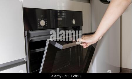 La main d'une femme ouvre la porte d'un four moderne dans la cuisine. Nettoyage catalytique Banque D'Images