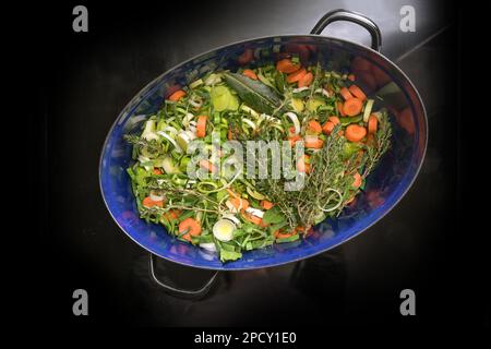 Légumes crus hachés comme les carottes, le céleri et le poireau avec des herbes comme le thym et les feuilles de Laurier dans une rôtissoire bleu foncé sur le dessus de poêle à induction noir, pour Banque D'Images