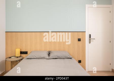 Lit double avec linge gris élégant, petite table de chevet et lattes décoratives en bois au mur. Concept de chambre à coucher compacte mais multifonction Banque D'Images