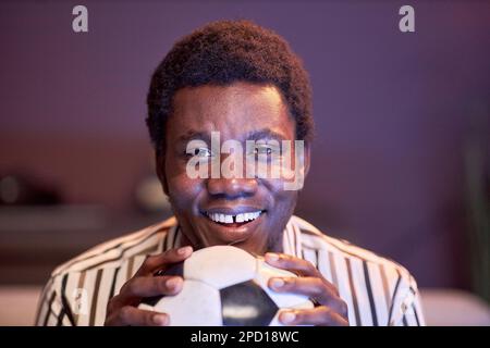 Gros plan d'un homme noir enthousiaste regardant un match de football à la télévision à la maison et tenant le ballon dans la lumière bleue Banque D'Images
