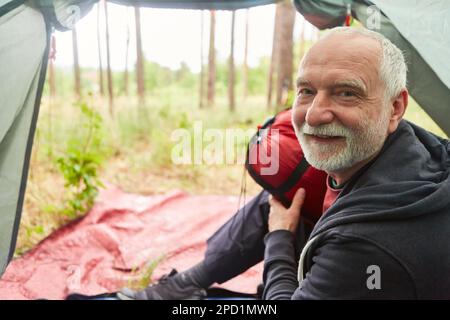 Portrait d'un homme âgé souriant assis avec sac à dos dans une tente pendant le camping en forêt Banque D'Images