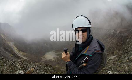 Jeune homme grimpeur marchant avec un manteau noir et un casque en utilisant le poteau de trekking seul par le cratère du volcan de Guagua Pichincha lors d'une journée nuageuse Banque D'Images