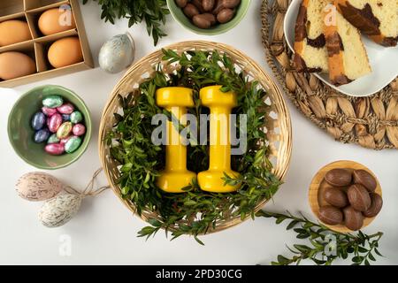 Haltères de gym dans le panier traditionnel en osier Pascal de Pâques. Concept de choix d'une alimentation saine. Plat de fitness avec bonbons au chocolat, gâteau, œufs, buis. Banque D'Images