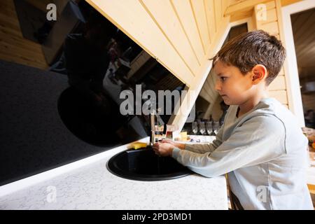 Le garçon lave les mains dans l'évier de la cuisine dans une petite cabane en bois confortable. La vie à la campagne. Banque D'Images