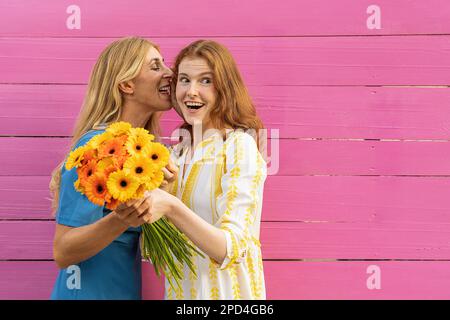 La petite amie murmure un secret dans l'oreille de son ami et tient un bouquet de fleurs de gerbera devant un fond de bois rose Banque D'Images