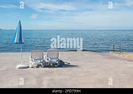 Automne avec des transats en plastique blanc vides à côté du parasol rayé bleu et blanc sur le littoral de la Croatie, Europe Banque D'Images