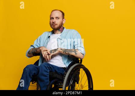 Un homme en fauteuil roulant a des problèmes avec le système musculo-squelettique regarde la caméra avec soin avec des tatouages sur ses bras assis sur un studio jaune Banque D'Images