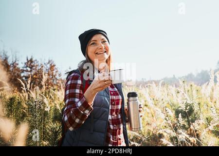 Femme souriante se détendant et appréciant le café pendant les vacances. Femme debout sur la piste et regardant loin tenant une tasse de café et thermos flacon. Banque D'Images